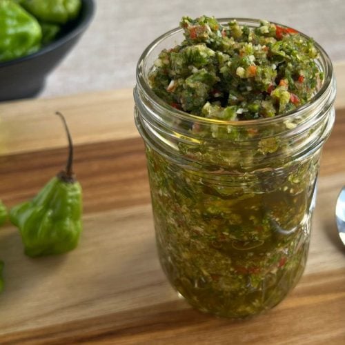A jar of sofrito sitting on a cutting board with a spoon and a few ahi dulce peppers