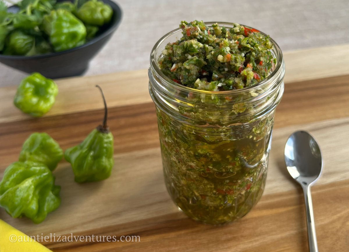 A jar of sofrito sitting on a cutting board with a spoon and a few ahi dulce peppers