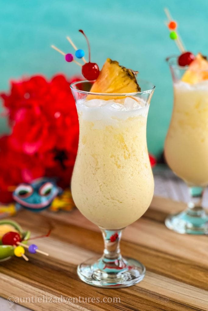 Pina Colada cocktail in a hurricane glass with pineapple wedge and cherry garnish sitting on a wood cutting board with flowers and garden lizard in the background