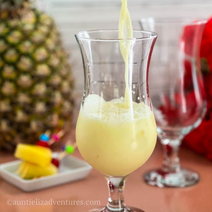 Pina colada cocktail being poured into a Hurricane glass which is sitting on a table with pineapple and pineapple cherry garnishes