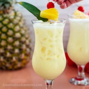 Pina colada cocktail in a Hurricane glass which is sitting on a table with pineapple with a hand placing the pineapple cherry garnishes on top of the cocktail