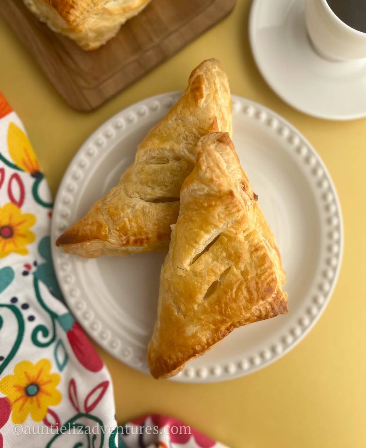 Two Guava Cream cheese pastries on a white plate surrounded by a cup of coffee, more guava cheese pastries on a wood board, and colorful floral napkin