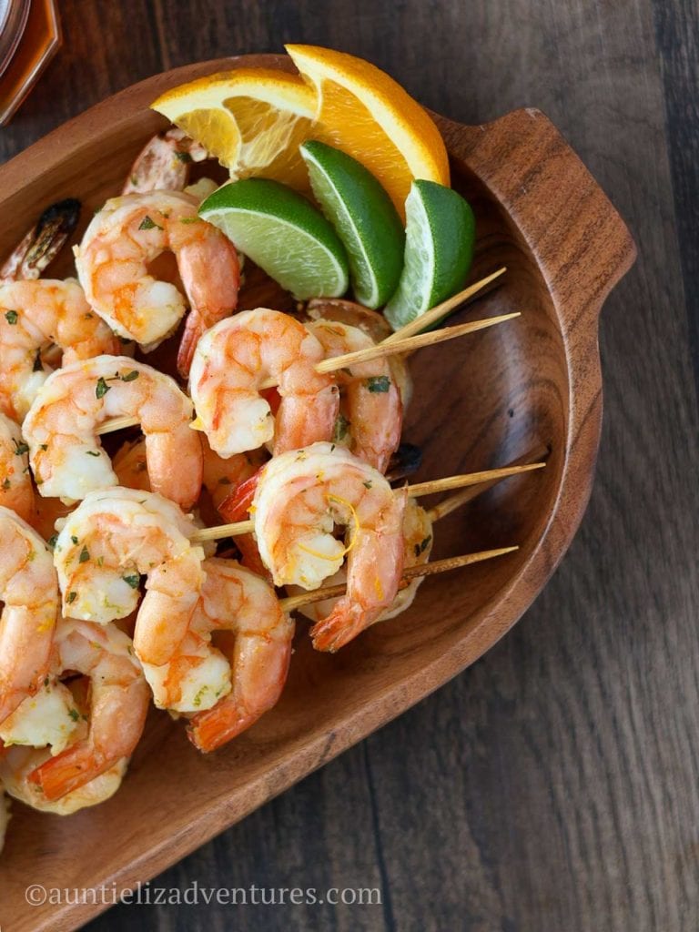 Wood table, with citrus marinated shrimp skewers, orange and lime slices on a wooden platter with a glass of beer beside the platter