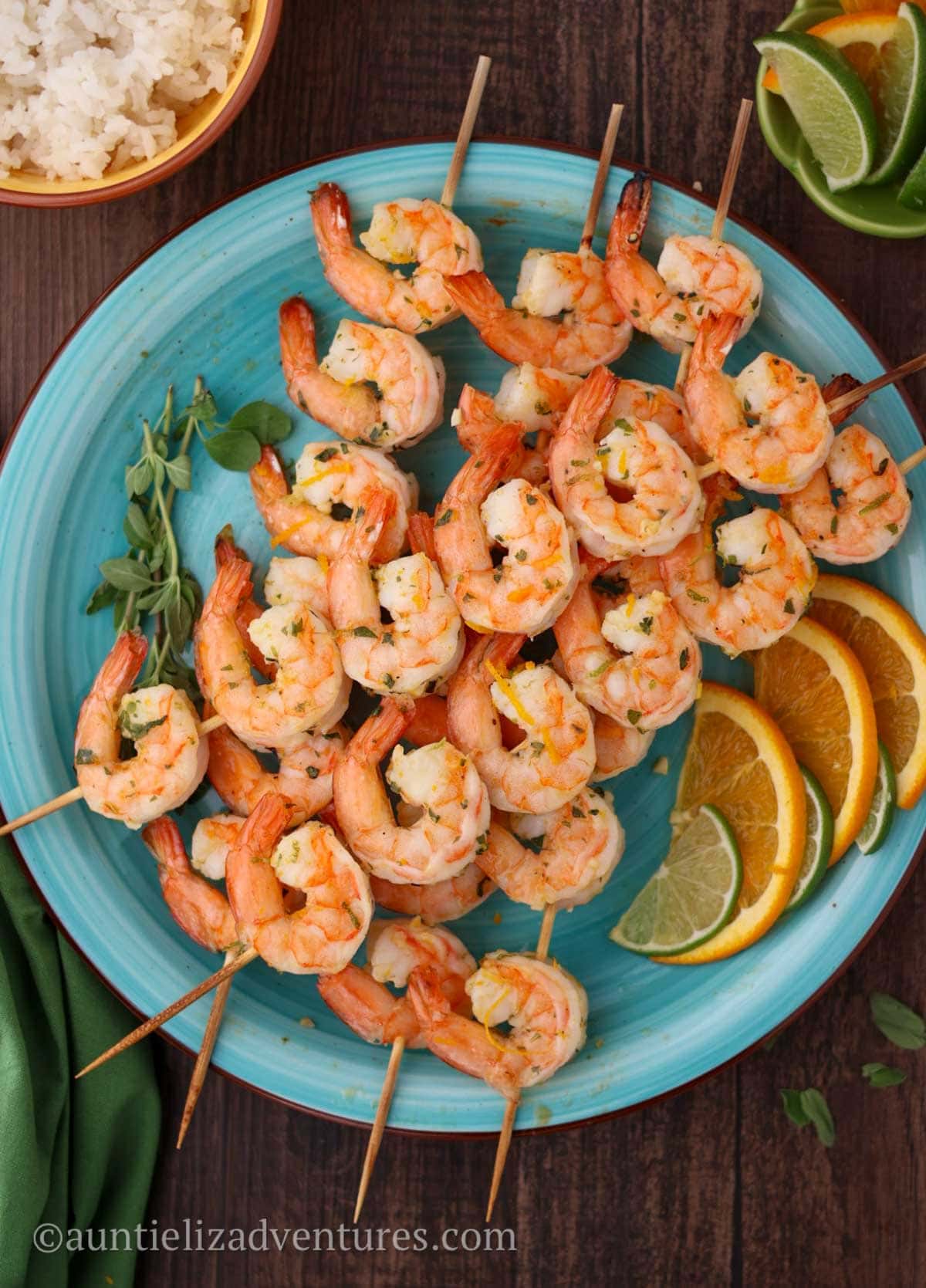 A wood table with an aqua plate of five cooked citrus marinated shrimp skewers garnished with oregano, orange and lime slices as a garnish. A green napkin, white rice in a yellow bowl, and lime and orange slices accompany the shrimp on the table.